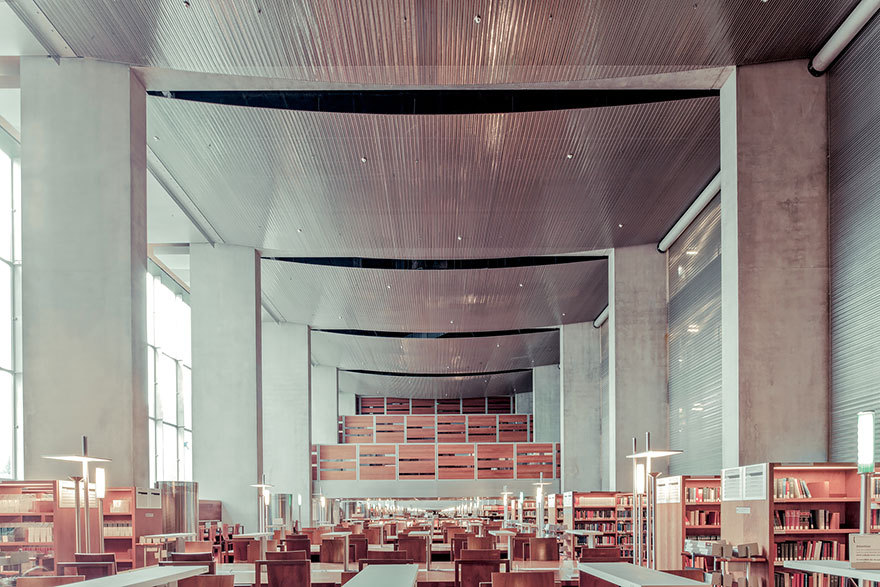 A modern library interior with concrete pillars, wooden shelving, rows of desks and warm lamps, conveying a quiet scholarly mood in muted grays and wood tones.